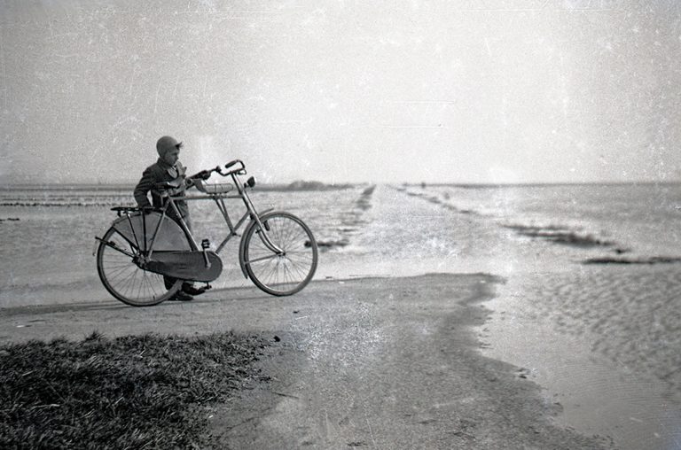 Zwart-wit foto uit de collectie van Johannes Boomsma en Johannes de Man waarop een jongen naast grote fiets uitkijkt over het bouwland dat onder water is gezet op Goeree-Overflakkee