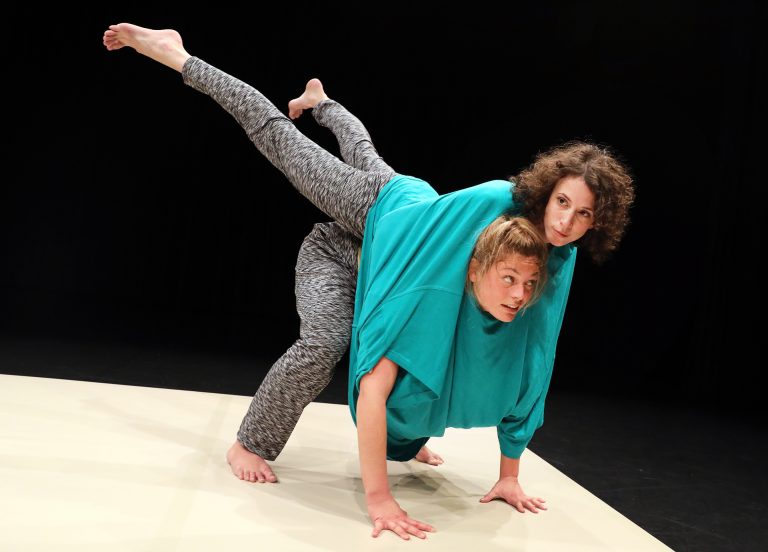 Twee dansers dragen allebei een zwartitte broek en zitten samen in een groot turquoise t-shirt terwijl de ene danser op de rug van de andere danser ligt