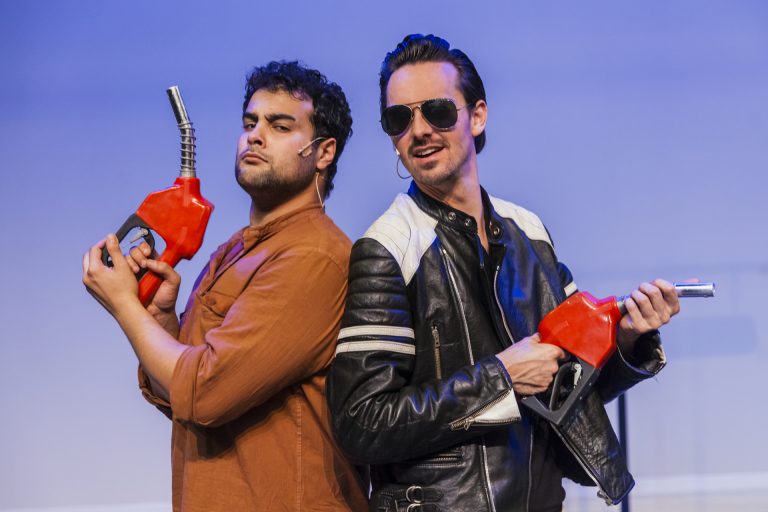 Twee mannen staan met de ruggen tegen elkaar en houden allebei een een brandstofpistool van een benzinepomp vast alsof ze twee cowboys met echte pistolen zijn