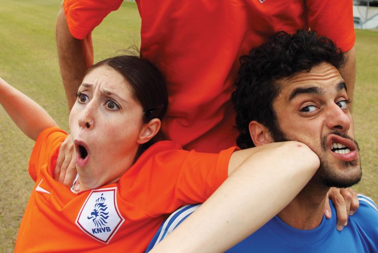 Actiemoment tijdens een voetbalwedstrijd waarbij een vrouw in een oranje shirt een elleboogstoot geeft aan een man in een blauw shirt terwijl iemand in een rood shirt bovenop hun springt