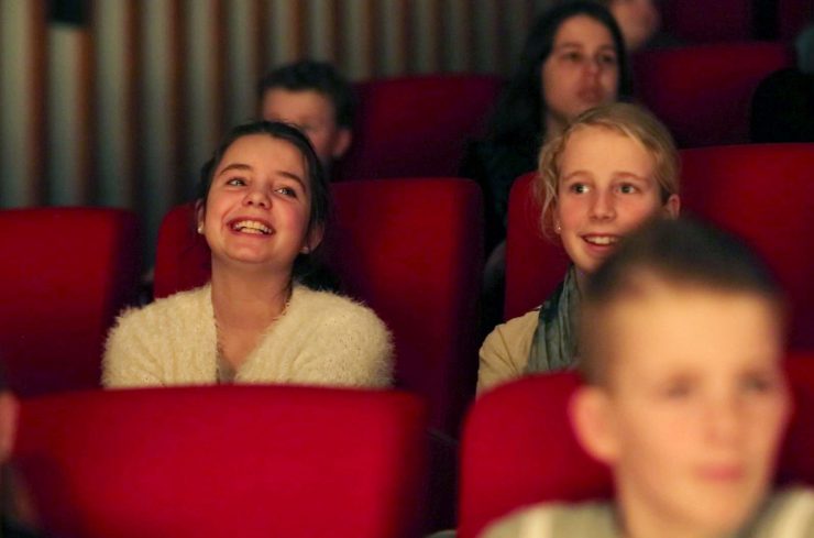 Leerlingen zitten op rode pluche stoelen in een theaterzaal en kijken naar een voorstelling