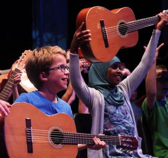 Meisje met hoofddoek houdt lachend een gitaar omhoog, naast haar zit en jongen met bril die ook een gitaar vasthoudt