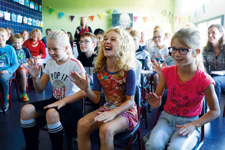 Leerlingen zitten op stoeltjes trommelen met hun handen op hun bovenbenen, het middelste meisje moet hard lachen