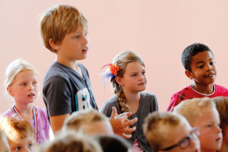 Jongen staat rechtop tussen een groepje zittende kinderen en geeft antwoord op een vraag