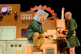 Scène uit de theatervoorstelling 'Wachten op Kado': een man in een beige broek en groene jas heeft over zijn hoofd een groot kado met een oranje strik erom. Hij zit op een grote machine, een andere acteur met een groene jas staat ernaast.