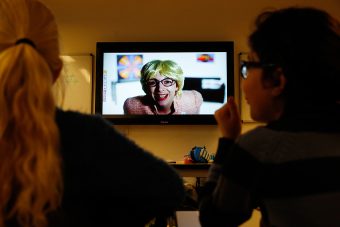 Twee leerlingen op de rug gezien terwijl ze kijken naar een film op het digibord met een mevrouw die hen toespreekt