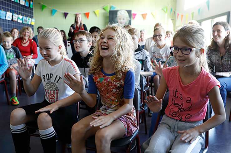 Leerlingen zitten op stoeltjes trommelen met hun handen op hun bovenbenen, het middelste meisje moet hard lachen