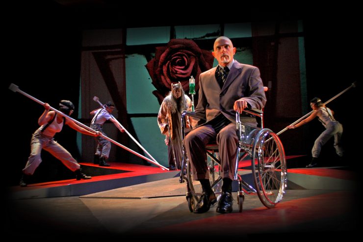Acteur met grijs pak en stropdas zit in rolstoel op podium, achter hem acteurs met lange stokken en een acteur met een infuus op een staander