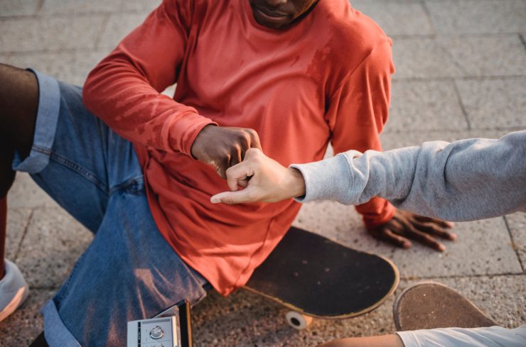 Jongen met rood shirt en korte spijkerbroek zit op zijn skateboard en geeft een boks aan een andere jongen in een grijze trui
