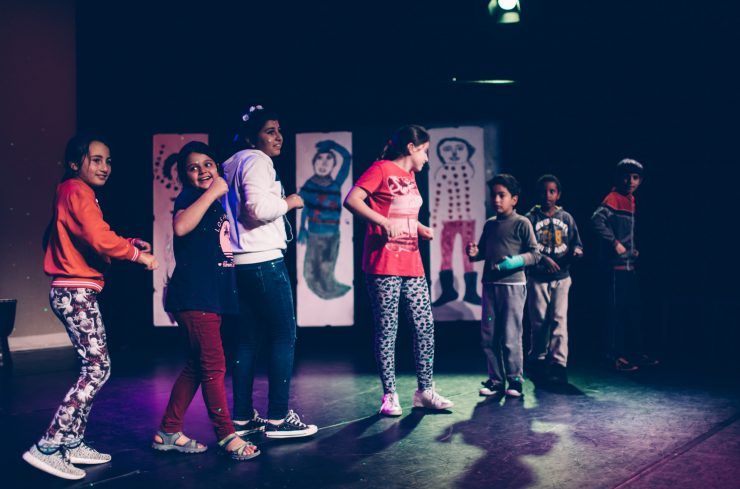 Zeven kinderen dansen op podium met als achtergrond grote kindertekeningen