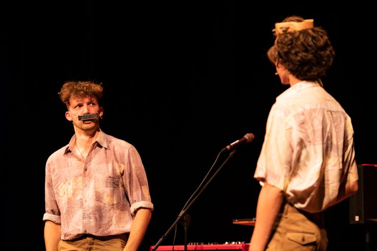 Twee mannen op toneel, de ene draagt een mondharmonica als snor en de andere heeft een tamboerijn op zijn hoofd