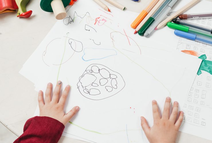 Handen van een kind rusten op een vel papier met een tekening daaromheen liggen stiften en potloden