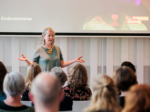 Vrouw in groene jurk en grote houten collier staat voor een groep docenten en geeft een presentatie