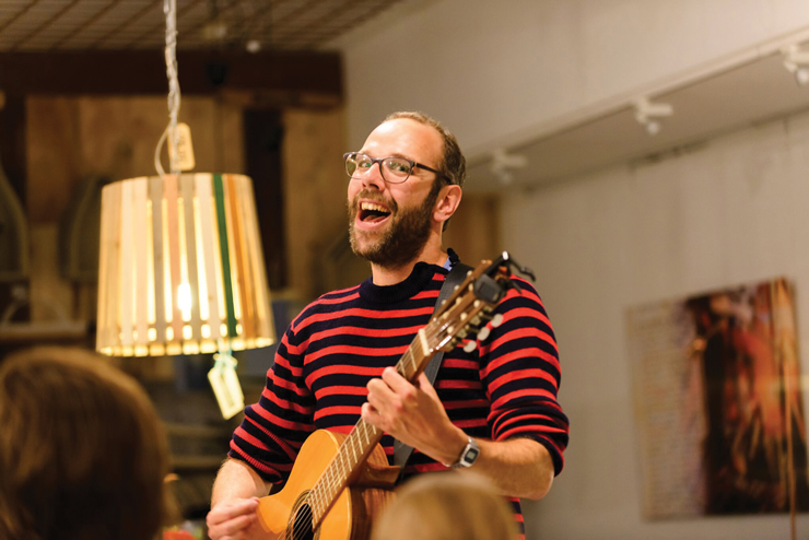 Man met baard en bril draagt zwart-rood gestreepte trui, lacht en speelt gitaar