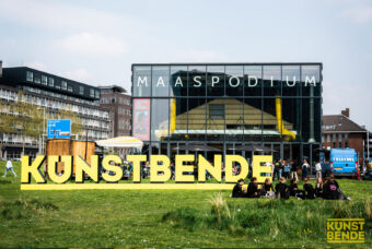 Het woord Kunstbende staat met grote gele letters in het gras. Op de achtergrond zie je het gebouw van Maas Podium.