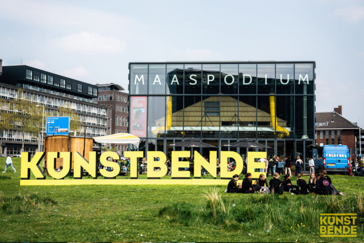 Het woord Kunstbende staat met grote gele letters in het gras. Op de achtergrond zie je het gebouw van Maas Podium.