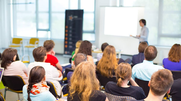 Een groep mensen gezeten op stoelen en van achteren bezien, luisterend naar een persoon die voor de groep staat
