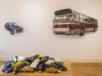 Tentoonstelling Te land, ter zee en in de lucht. Een auto en bus aan de muur. Gebruikte kunstzinnige flessen op de grond.