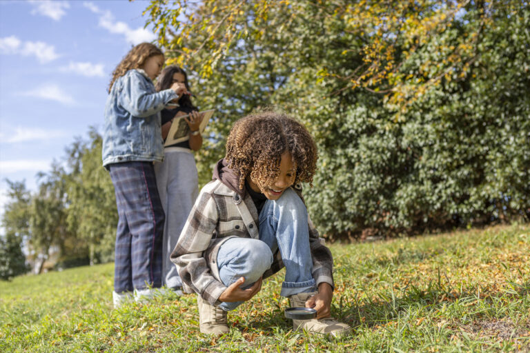 Erfgoed in de klas Jongetje zit met vergrootglas in het gras. 2 kinderen staan in de achtergrond.